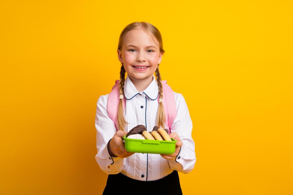 Young schoolgirl with pigtails holding a lunchbox filled with snacks.