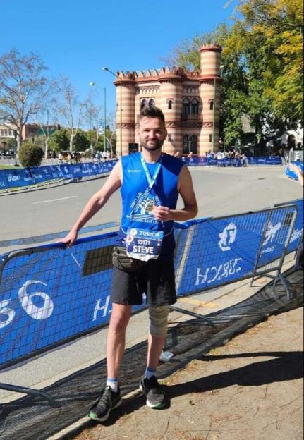 Steve Roe, wearing a blue running shirt with a medal around his neck and a leg brace, stands next to a barrier after a race.