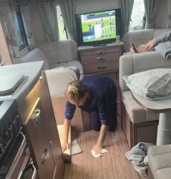 A woman kneeling and wiping the floor inside a camper van, while another person watches TV in the background.