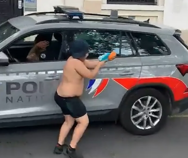 A shirtless boy aiming a water pistol at a person inside a French police car.