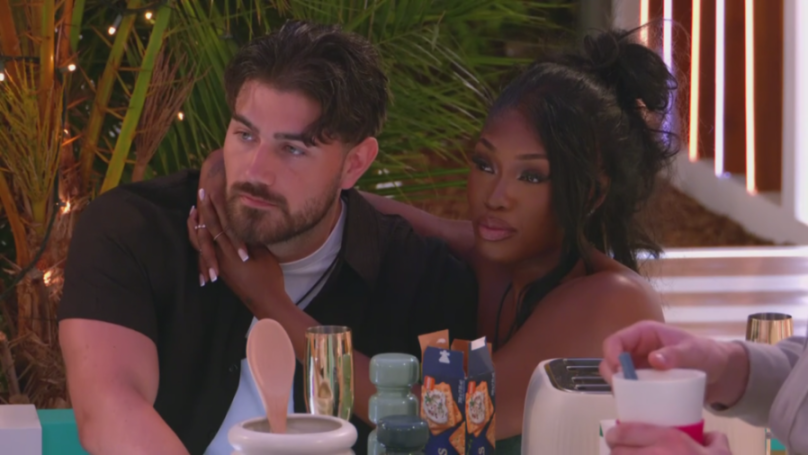 A man with a beard and dark hair sits next to a woman with long dark hair who has her arm around his neck, looking at him.