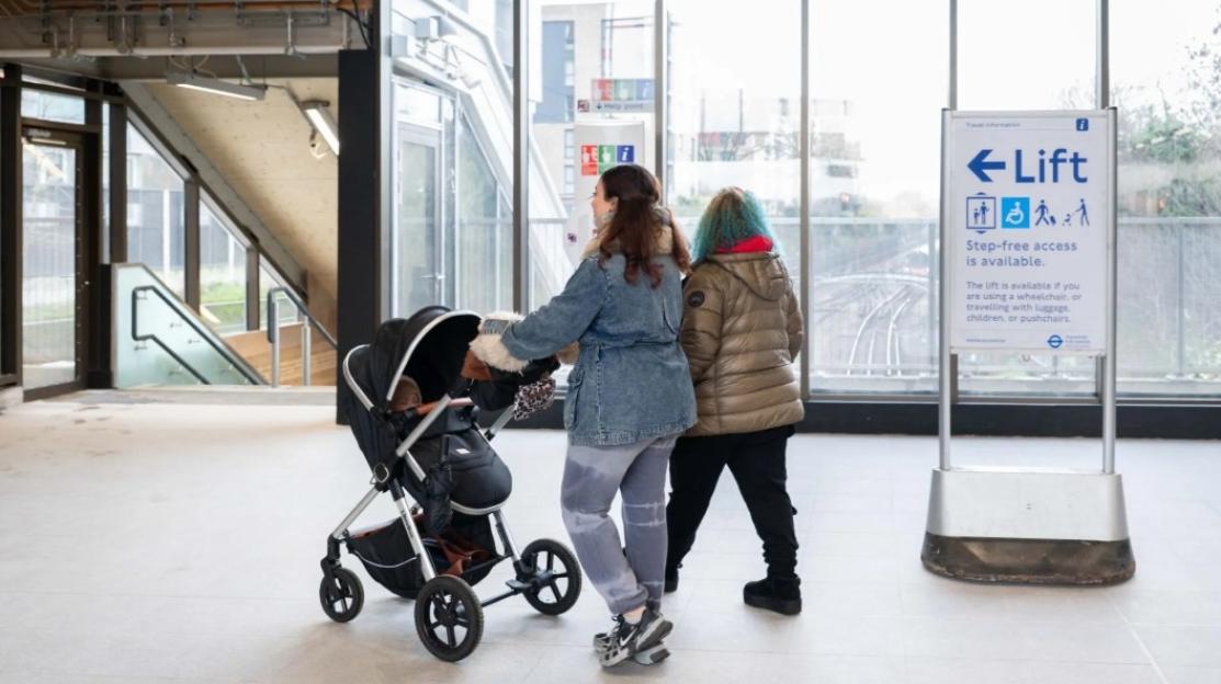 Two women with a baby in a stroller standing near a sign for step-free lift access at Colindale station.