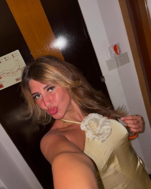 A young woman with long blonde hair and plump lips, wearing a light yellow top with a large white flower, making a kissing face at the camera.
