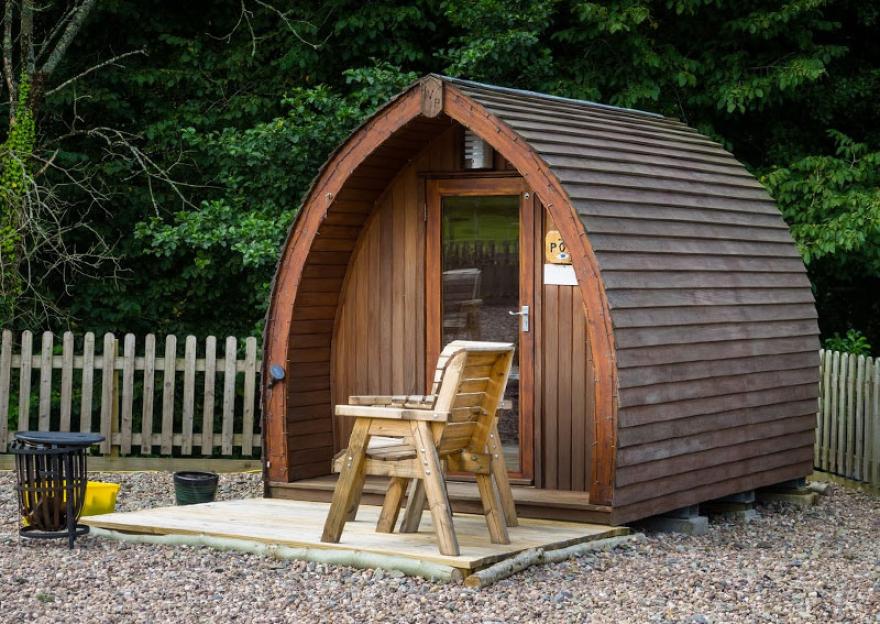 Wooden camping pod with a small porch, chair, and fire pit in front of a wooden fence and trees.