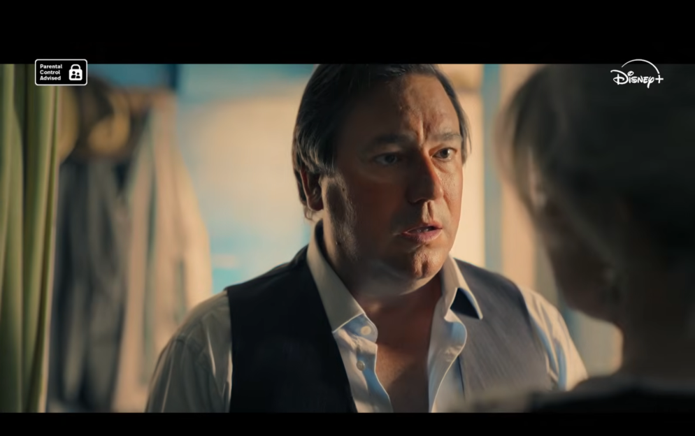 A man in a white shirt and dark vest looks concerned as he talks to someone out of frame.