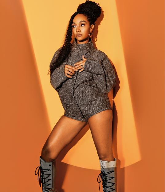 Leigh-Anne Pinnock poses in a tweed romper and white lace-up boots against an orange background.