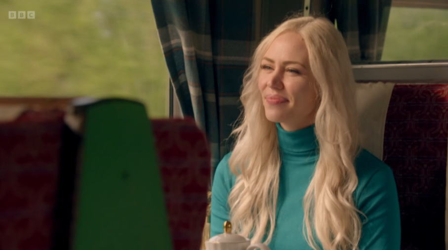 Ellie from Traitors, with long blonde hair and a blue turtleneck, looking to her left.