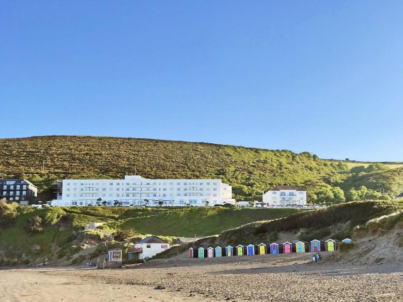 Tarka Holiday Park Devon with a long white building and colorful beach huts by a sandy beach.