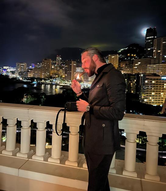 Ricky Lawrence lights a cigar on a balcony overlooking a city at night.