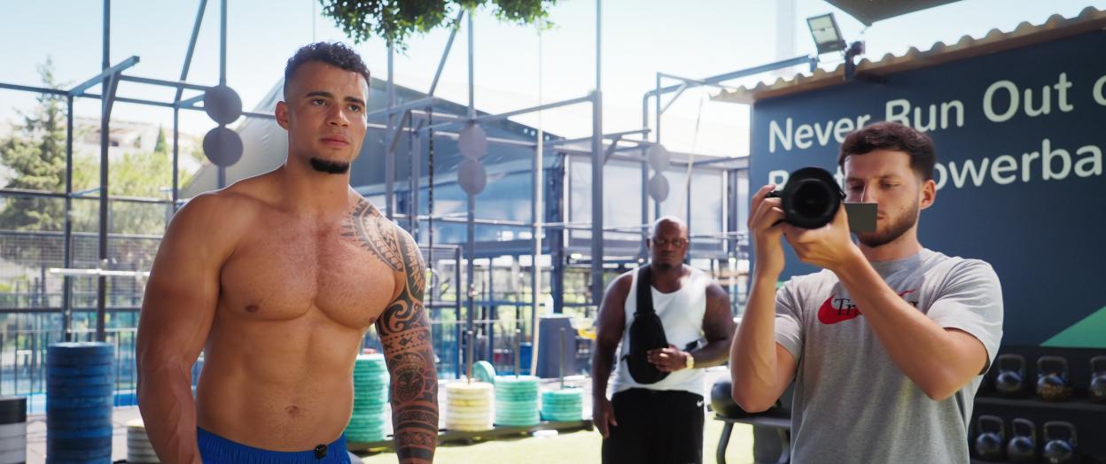 Harrison Sullivan, shirtless and tattooed, in a gym, with a cameraman filming him.