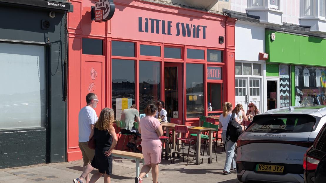 People walk past the Little Swift bar and wine shop, with outdoor seating, and a parked car.