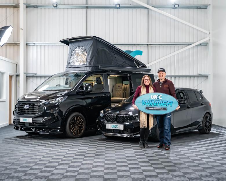 A man and a woman standing in front of a black camper van and a black car, holding a blue sign that says "UKC WINNER".