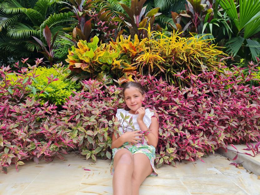 A young girl smiling and sitting on a tiled surface in front of a colorful backdrop of lush plants and foliage.