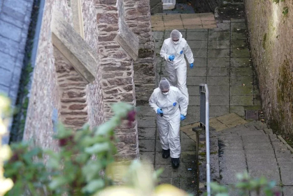 Forensic investigators in protective white suits walking down a stone pathway near a building.