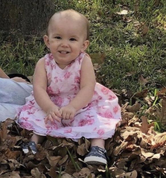 Annie, a toddler wearing a pink floral dress, smiles while sitting in a pile of autumn leaves.