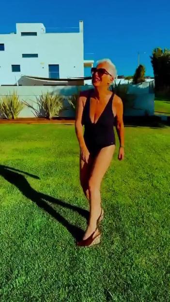 Denise Welch in a black swimsuit, sunglasses, and wedge sandals, walking on green grass in front of a white house.