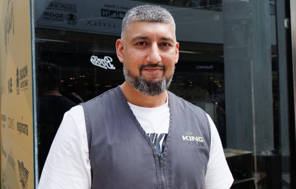 A man with gray hair and beard, wearing a white shirt and gray vest, stands in front of a storefront with a "King Solis" logo on his vest.