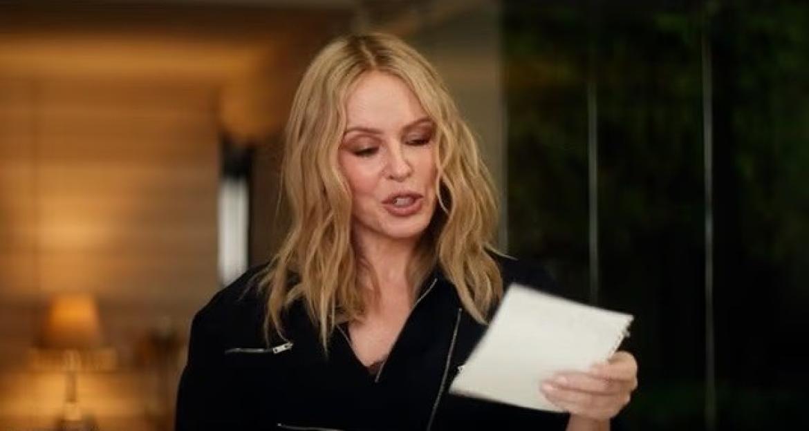 A woman with long wavy blonde hair wears a black jacket while reading from a white sheet of paper.