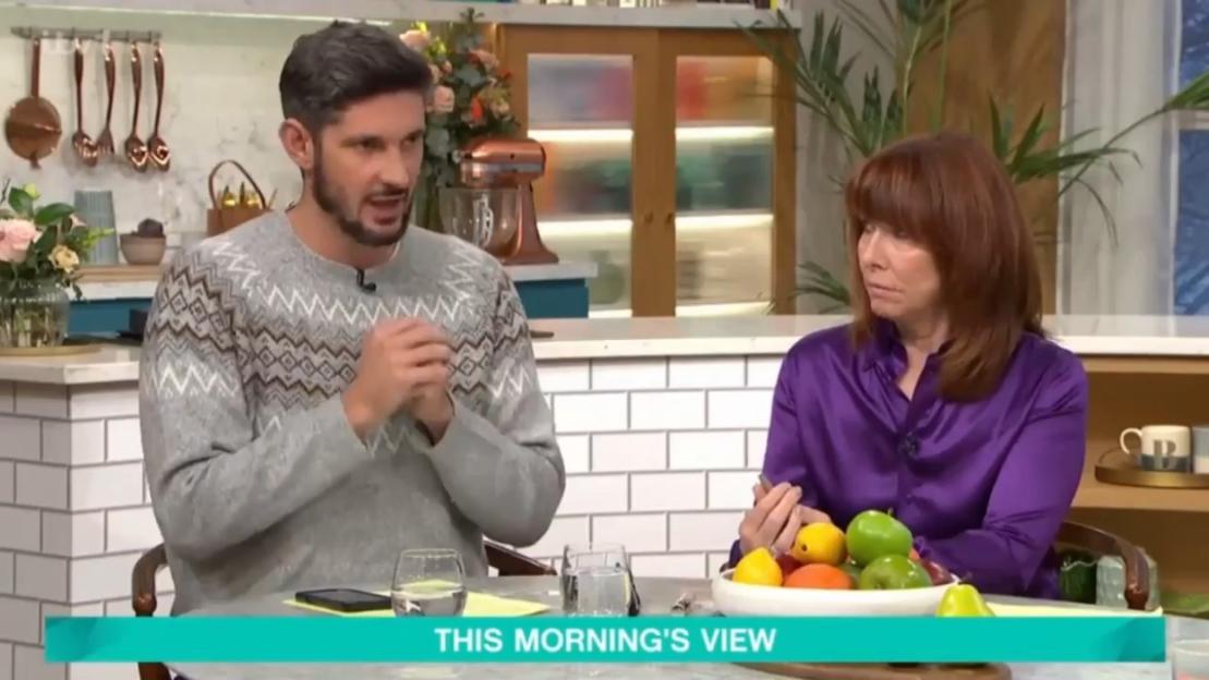 A man in a patterned sweater talking to a woman in a purple shirt on a TV show set.