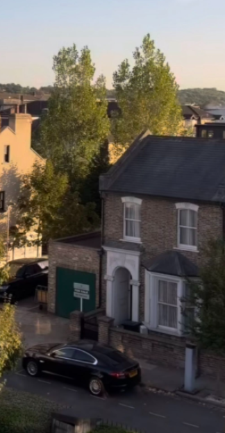 A black car parked in front of a brick house with a "For Sale" sign in the yard.