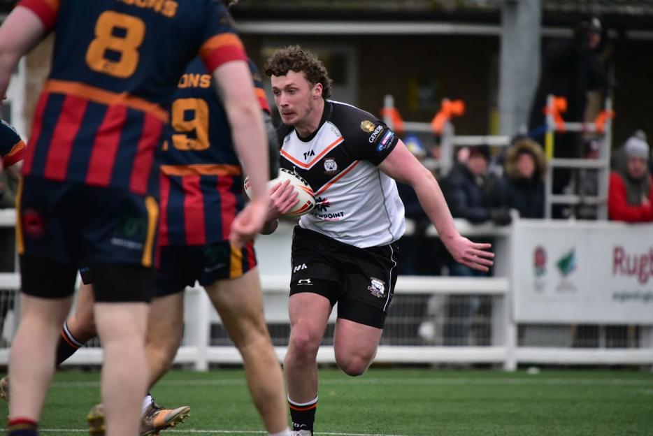 A rugby player in a white and black jersey holding the ball while running on a green field.