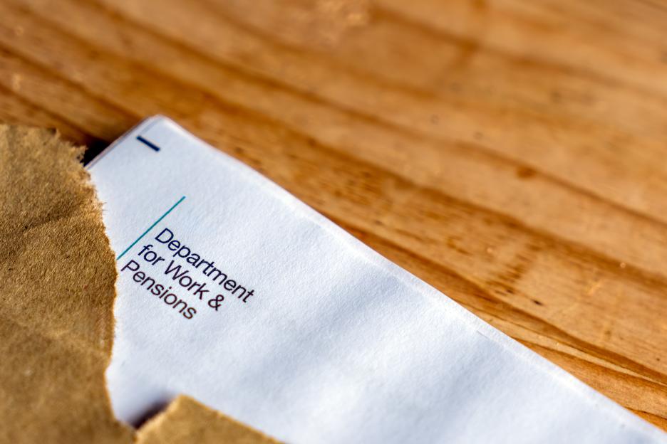 A brown, torn envelope on a wooden table partially reveals a white letter from the "Department for Work & Pensions."