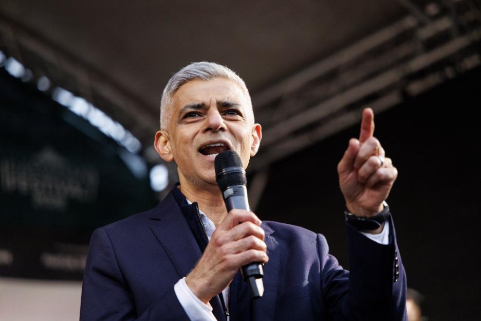 Sadiq Khan speaking into a microphone, with his right hand raised and index finger pointing up.