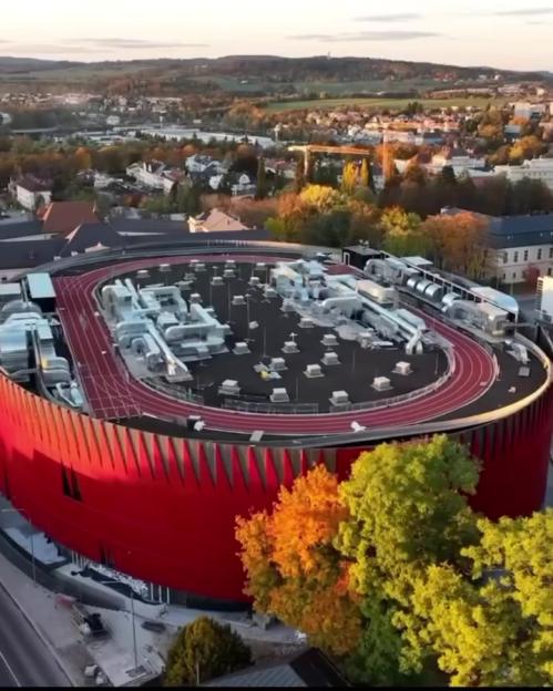 A red cylindrical stadium with a running track on its roof, surrounded by trees and a town in the background.