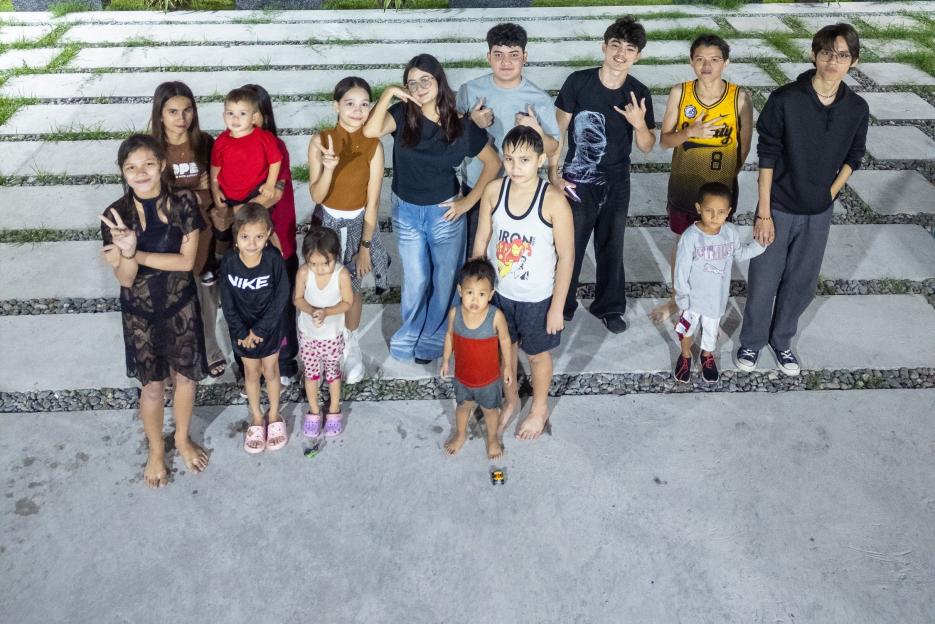 Large group of people, including adults and children, gathered in an outdoor area at night with a building and swimming pool in the background.