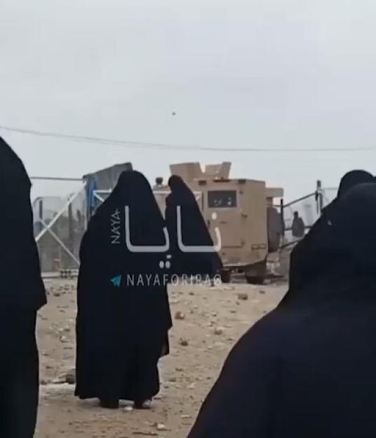 Women in black hijabs and abayas, possibly ISIS brides, in a camp with a military vehicle in the background.
