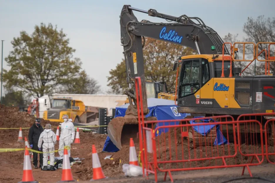 Construction site with an excavator, two workers in white hazmat suits, and one worker in a black jacket, standing next to an area covered by a blue tarp.