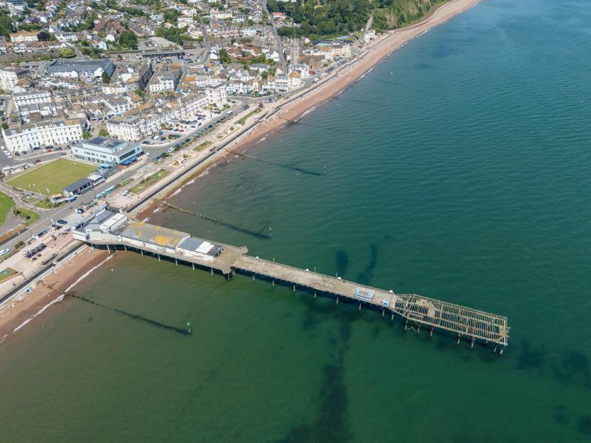 Aerial view of Teignmouth's partially dilapidated pier extending into the sea, with the town and beach visible on the left.