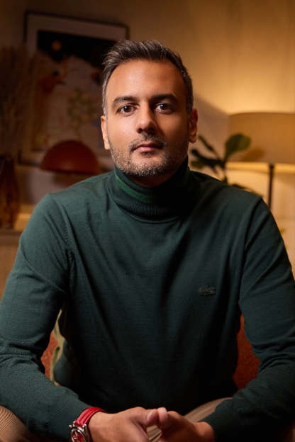 Dr. Syed, a man with a beard, wearing a green turtleneck and a red-banded watch, looks at the camera.
