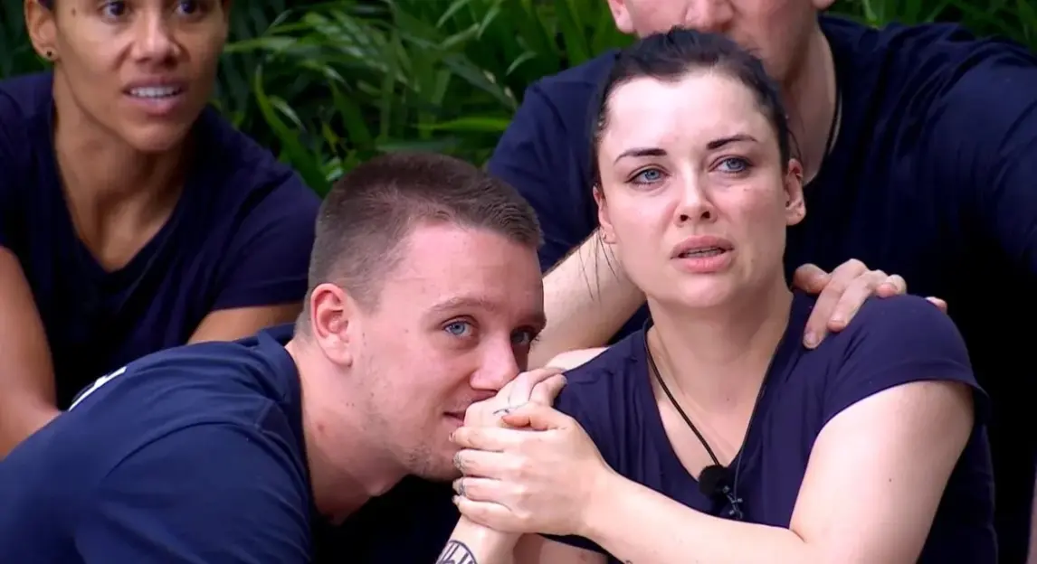 A man with blue eyes comforts a woman with dark hair who appears distressed, holding her hand and resting his head near her shoulder.