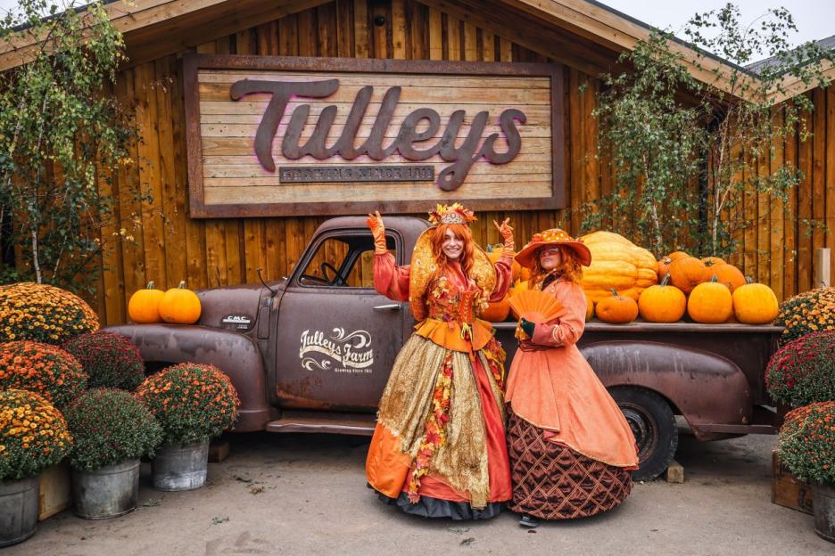 Two women in elaborate orange and gold dresses and hats stand in front of a rustic farm setting with pumpkins, chrysanthemums, and an old truck.