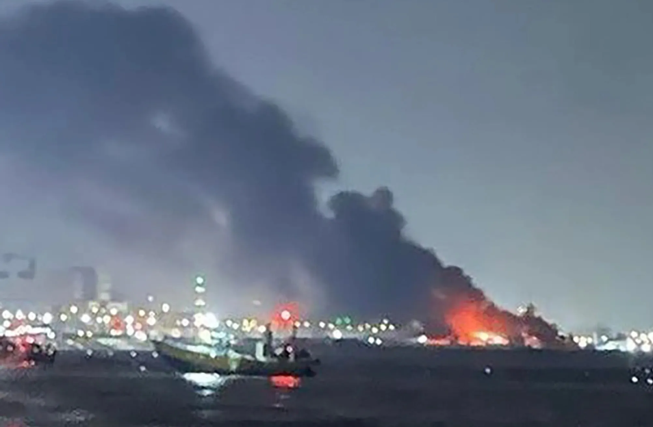 A fire at night over water with a large plume of smoke and distant city lights.