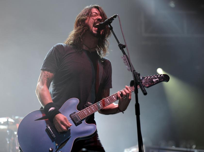 Dave Grohl of Foo Fighters playing a blue Gibson guitar and singing into a microphone on stage.