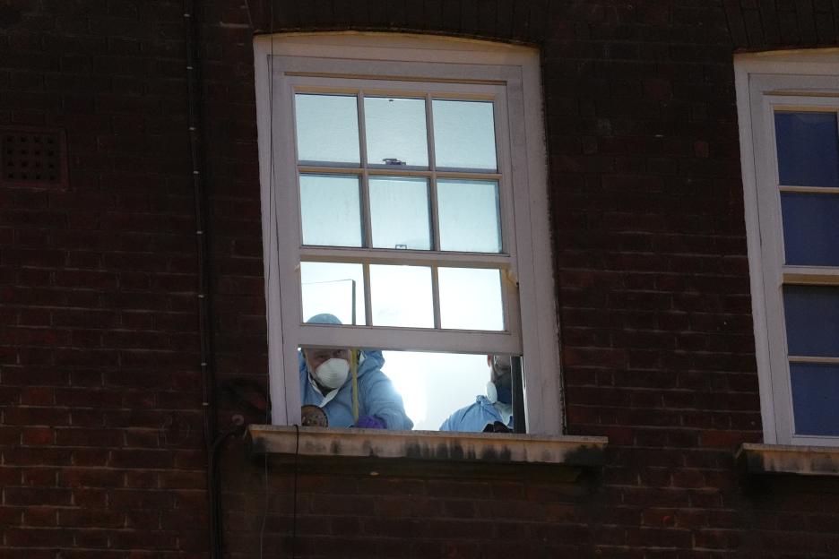 A forensic investigator wearing a white mask and blue protective suit looks out a window.