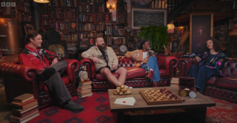 Four people sit on red leather couches in a book-filled room, engaging in conversation around a coffee table with a chess set.