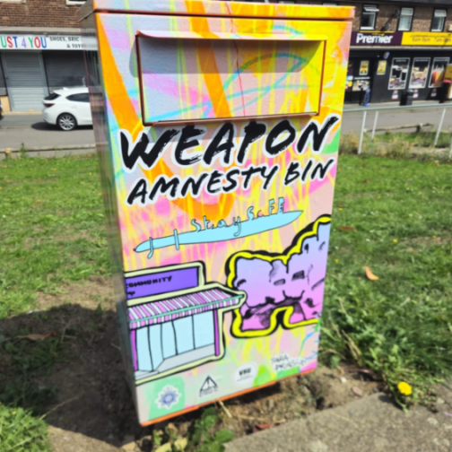 A brightly colored "Weapon Amnesty Bin" with graffiti art on it, placed outdoors on grass next to a sidewalk.