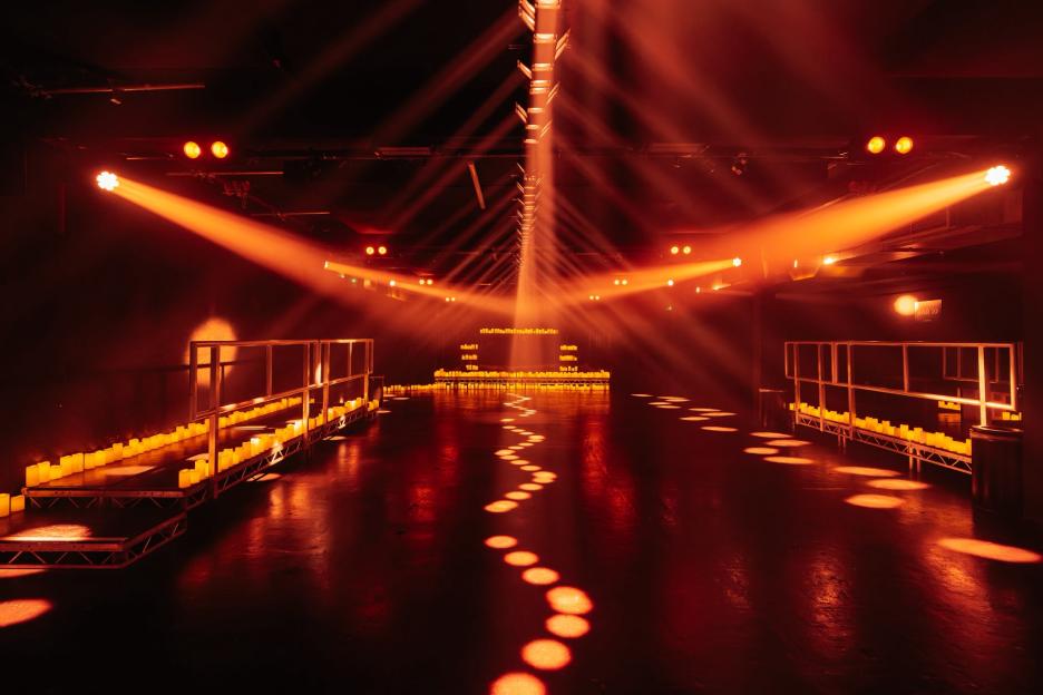 An immersive exhibition space illuminated with warm orange spotlights, candles, and a dotted light path on the floor.