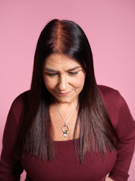 Woman with noticeable hair loss along her part, contrasting with her darker hair, and wearing a maroon ribbed top and a silver necklace, looking downwards against a pink background.