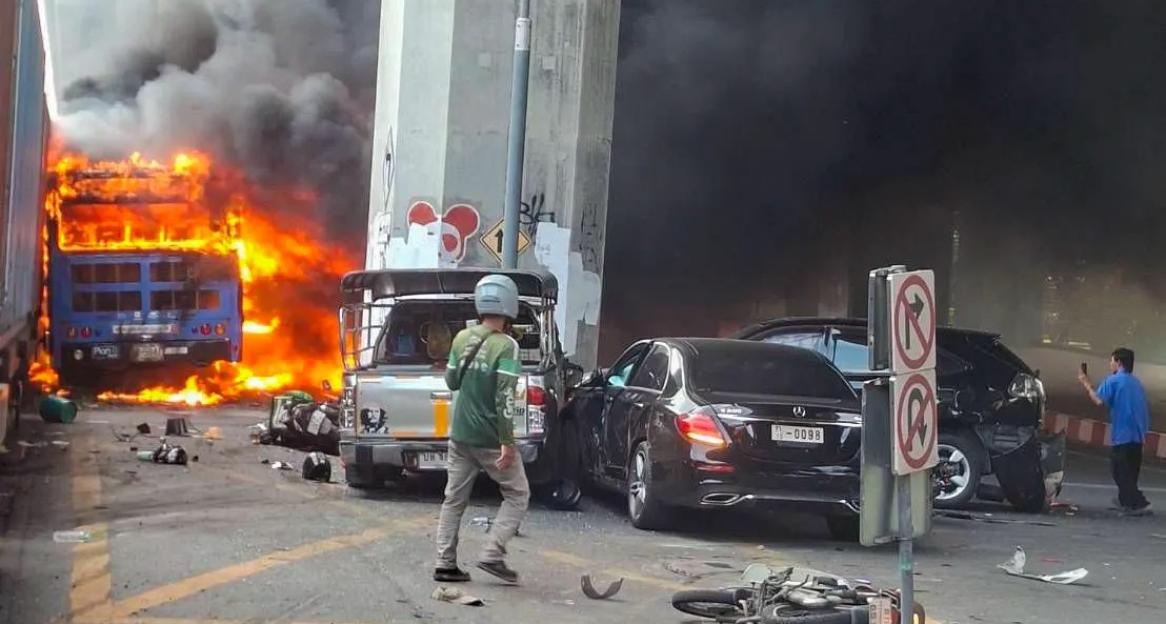 A bus engulfed in flames with dark smoke billowing into the sky after a collision with a train in Bangkok, with wrecked cars and debris scattered nearby.