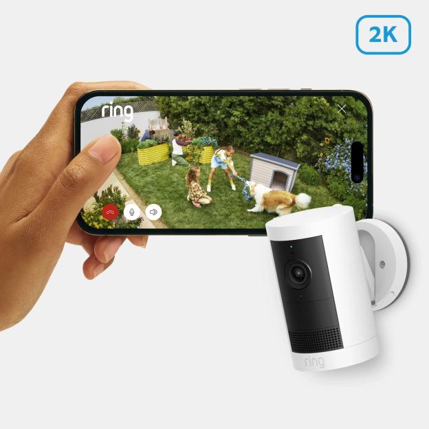 A hand holding a smartphone displaying a Ring camera's 2K video feed of a backyard with children and a dog, next to a white Ring camera.