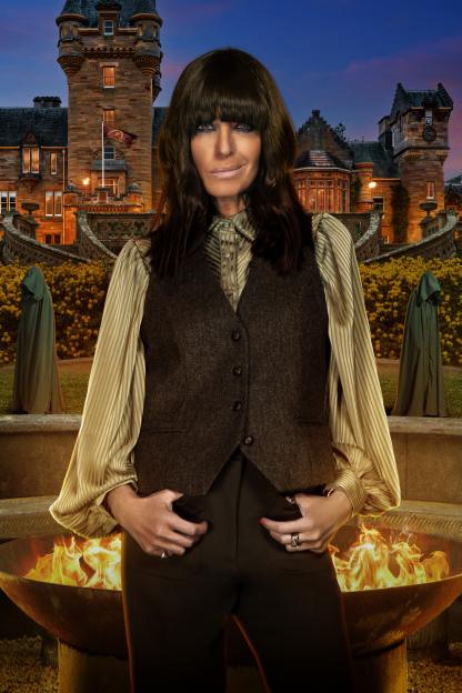 Claudia Winkleman posing in a dark vest and striped shirt, in front of a castle and a fire pit, with two hooded figures in the background.
