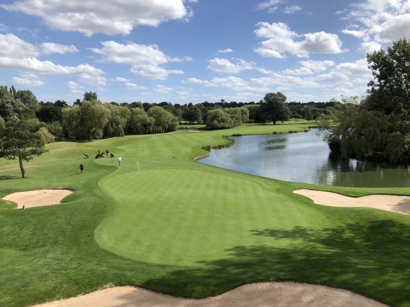 The Brabazon golf course features a green fairway with sand bunkers and a lake, under a blue sky with white clouds.