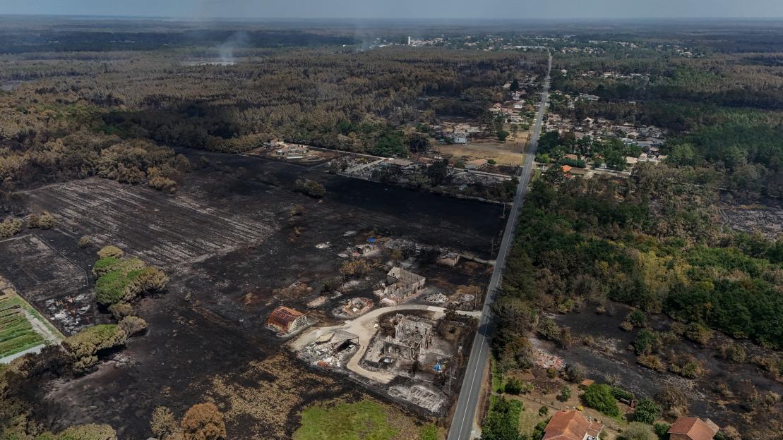The remains of a stone building, a burnt field, and a destroyed car after a wildfire.