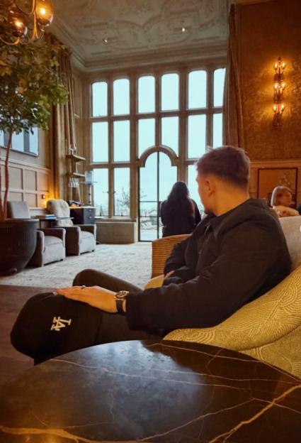 A person in a black jacket and a baseball cap on their lap, looking away in a large room with tall windows.