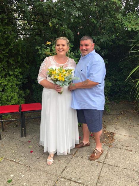A smiling couple stands outdoors with the woman in a white dress and holding a yellow bouquet, and the man in a blue shirt and shorts.