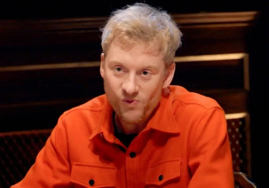 A photo of comedian James Acaster wearing an orange jacket on The Traitors.